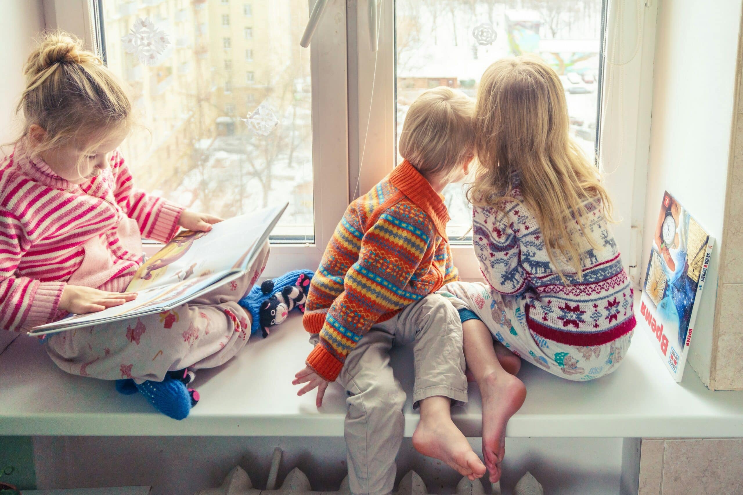 Three children sat on a windowsill, reading and looking out of the window